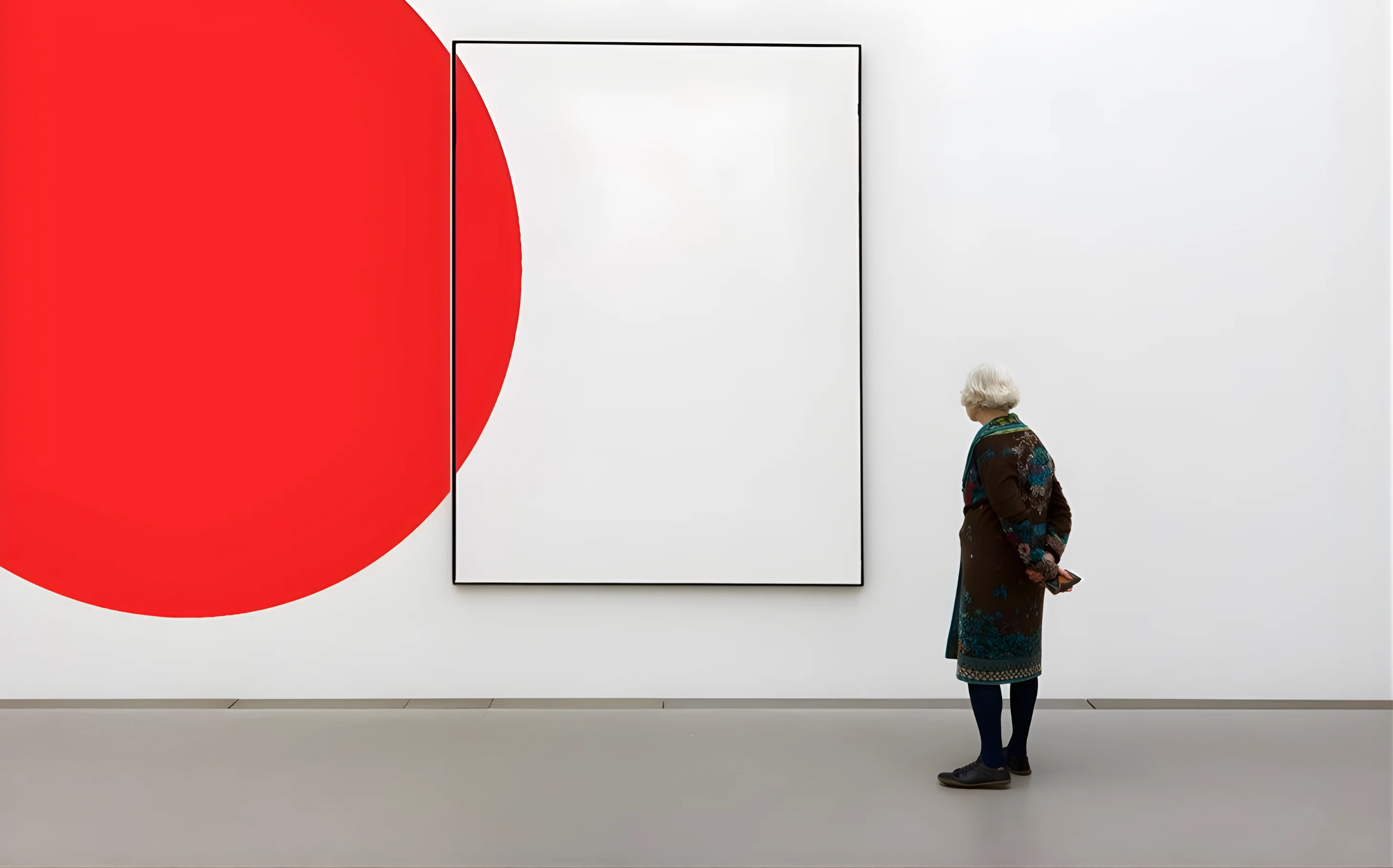 Older woman with white hair and patterned coat observing minimalist white framed artwork next to a large red circle painted on a white gallery wall.