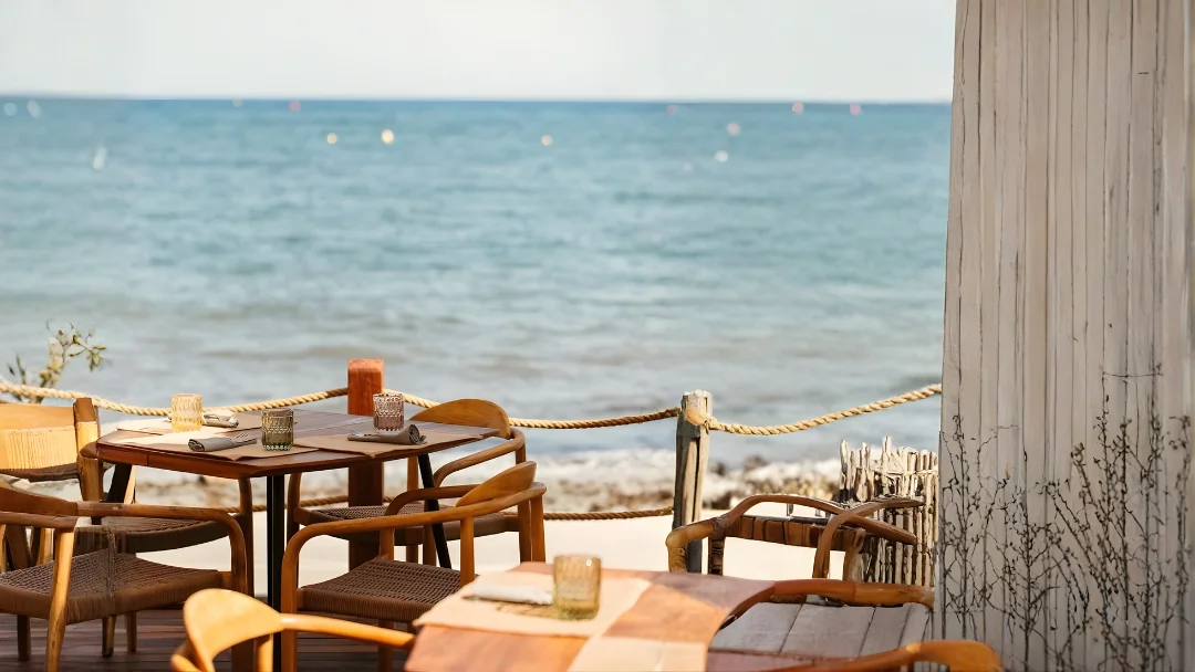 Outdoor wooden tables and chairs set for dining near a beach with the ocean in the background.