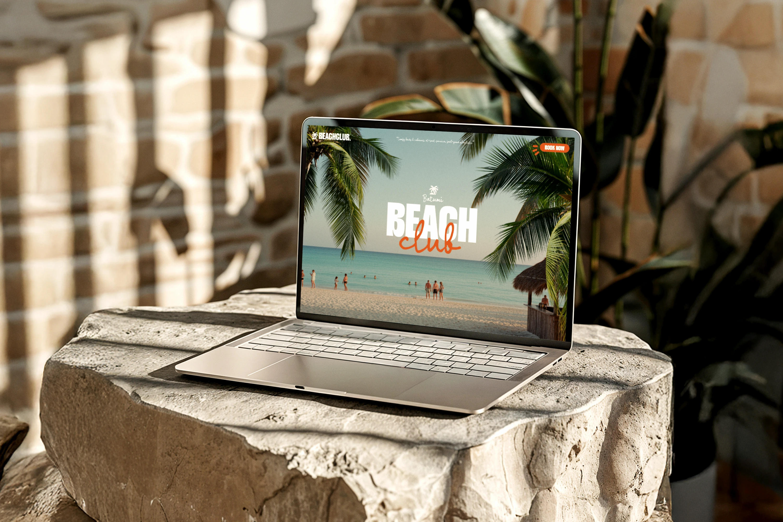 Laptop on a stone table displaying a beach club website with palm trees, ocean, and people on the shore.