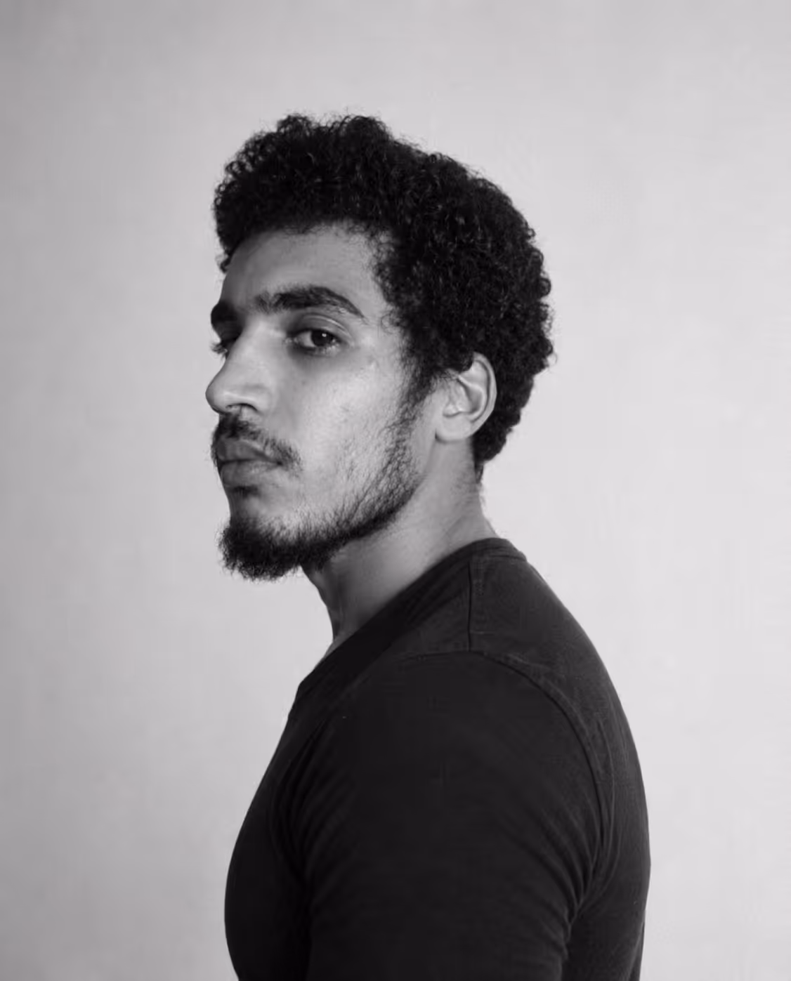 Homme à cheveux bouclés et barbe courte en profil, regardant vers la caméra, fond neutre.