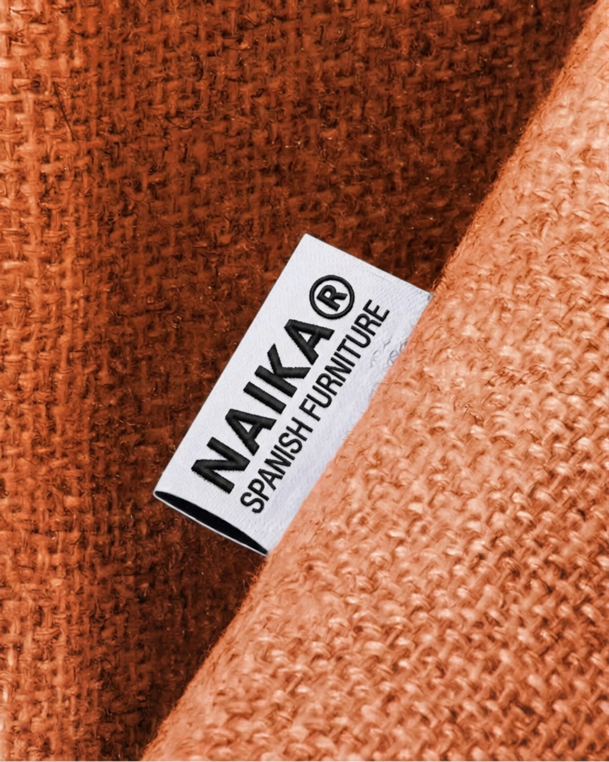 Étiquette blanche avec inscription 'NAIKA SPANISH FURNITURE' cousue sur un tissu texturé orange.