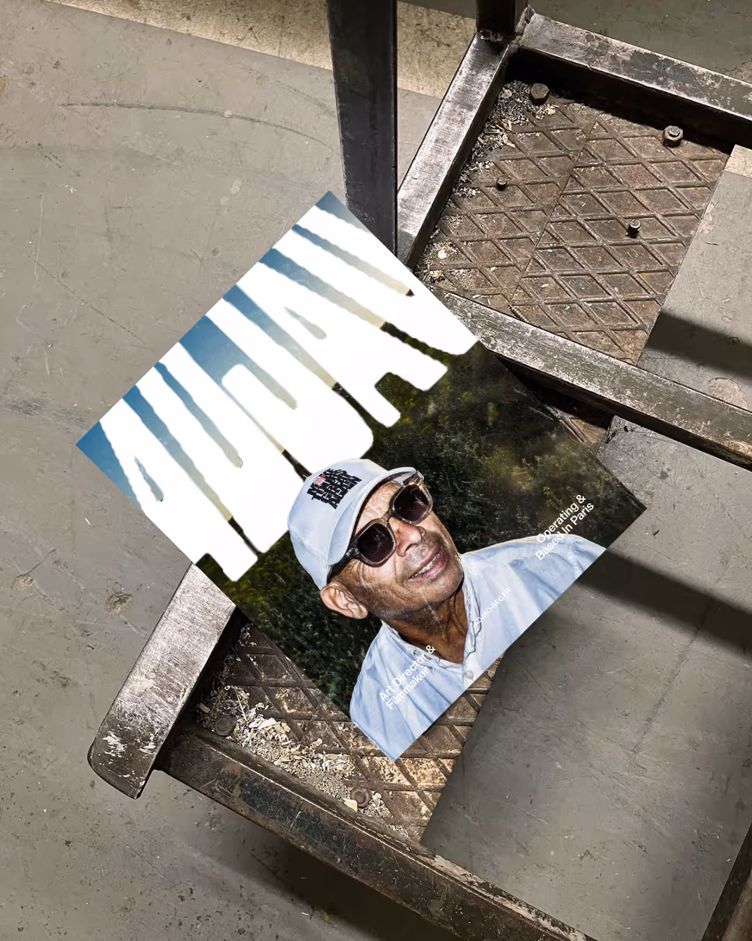Magazine with a man wearing glasses and cap on a metal industrial chair with textured dirt floor.