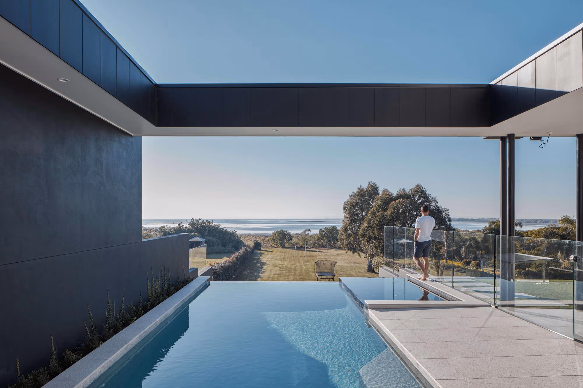 Modern house with an infinity pool overlooking a lawn, trees, and water, with a man standing near glass railing.